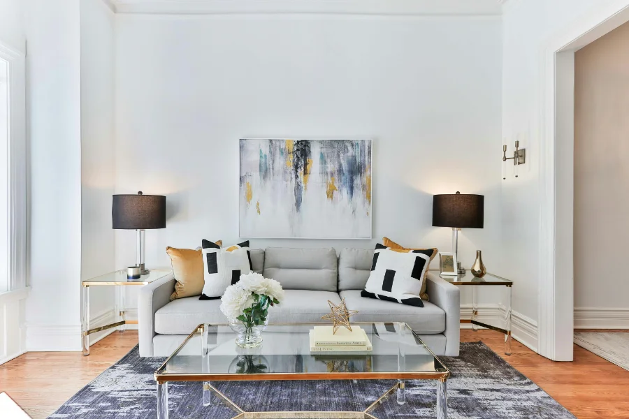 A bold living space with a blue area rug, grey couch, black, white, and gold pillows, accents, and a painting with the same colours sits on the white walls