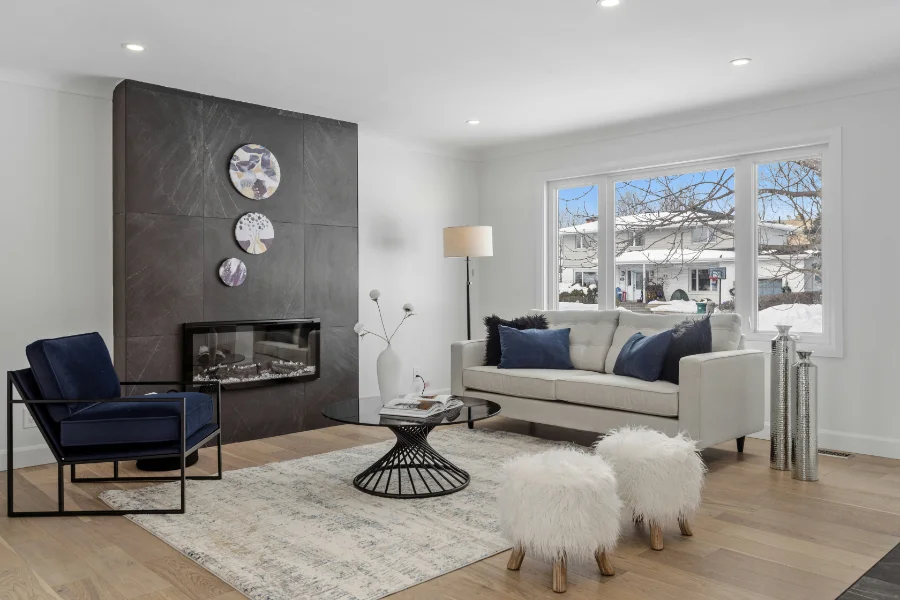A modern living room with a black slab fireplace, fuzzy white stools, navy blue armchair, and grey couch with navy blue pillows.