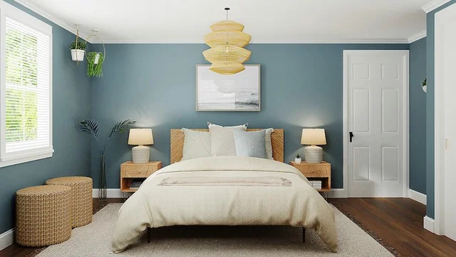 A bed is focus center, with light beige linens, and light coloured pillows, while the walls are painted in a med-light blue.